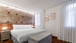 A bedroom with a brick wall, a bed with white linens, a nightstand with a lamp, and a large window with sheer curtains. - SAN SILVESTRO HOUSE VENICE (Venezia)