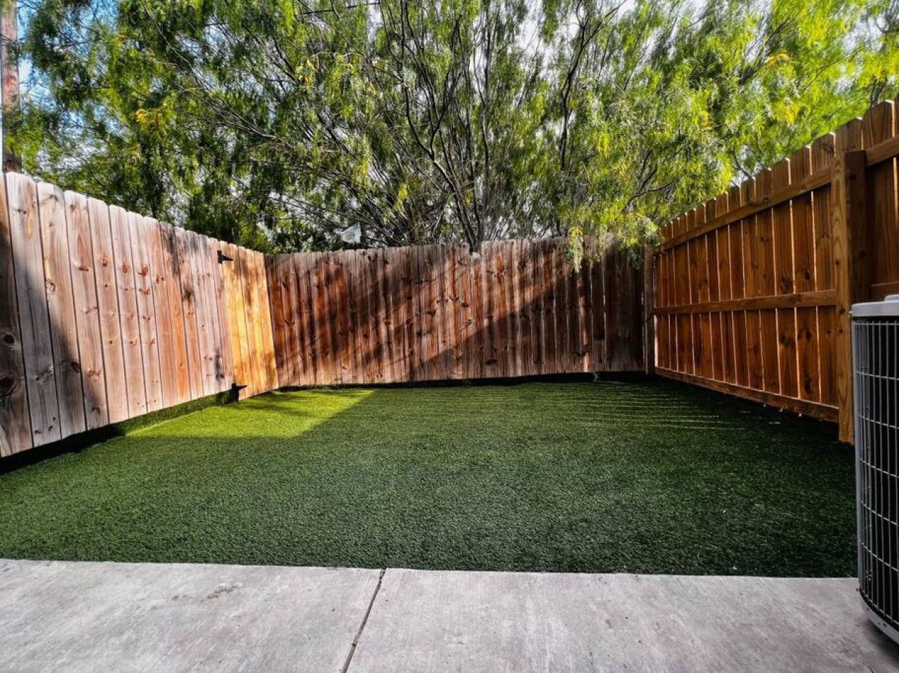 Un patio trasero con césped artificial verde, cerca de madera y patio de concreto.