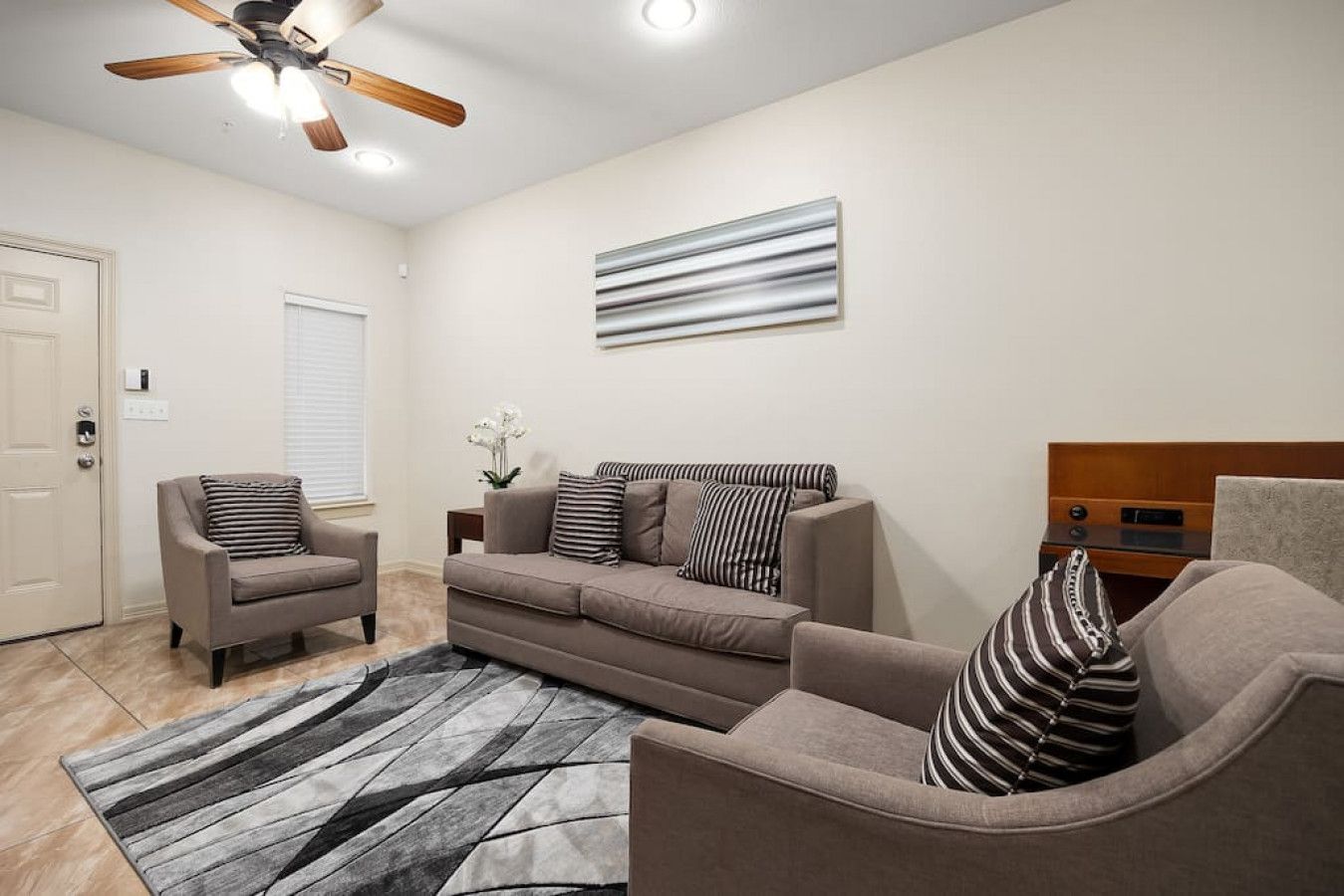 A living room with a sofa, armchairs, a coffee table, a rug, a ceiling fan, and a wall-mounted artwork.