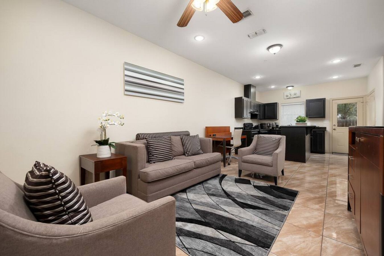 Ein modernes Wohnzimmer mit einer Couch, Sesseln, einem Couchtisch und einem Teppich. Im Hintergrund ist eine Küche mit einem Fernseher und Schränken zu sehen.