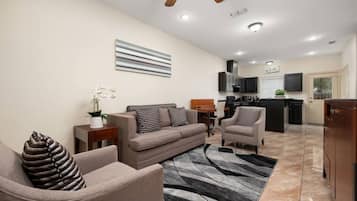 Ein modernes Wohnzimmer mit einer Couch, Sesseln, einem Couchtisch und einem Teppich. Im Hintergrund ist eine Küche mit einem Fernseher und Schränken zu sehen.