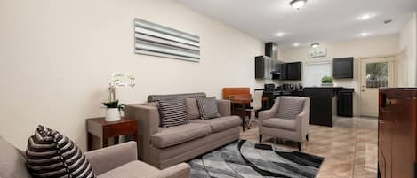 A modern living room with a sofa, armchairs, a coffee table, and a rug. There is a kitchen in the background with a TV and cabinets.