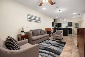 A modern living room with a sofa, armchairs, a coffee table, and a rug. There is a kitchen in the background with a TV and cabinets. - Nest at Hummingbird 3 w King bed free parking  Patio (Pharr)