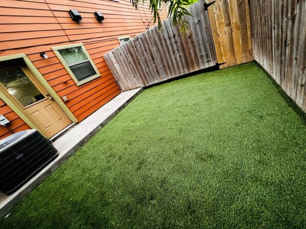 A backyard with artificial green turf, a wooden fence, and a building with orange siding and a door.