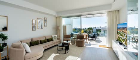 A modern living room with a curved sofa, a coffee table, and a TV on a stand. There is a balcony with a view of the city.
