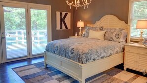 Ein Schlafzimmer mit einem großen Bett, einer Chandelüster, einem Buchstaben 'K' an der Wand und einem Fenster mit Jalousien.
