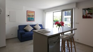 Un espace de vie compact comprenant un coin cuisine, un canapé bleu, un coin repas avec des tabourets de bar et une grande fenêtre donnant sur l’extérieur.