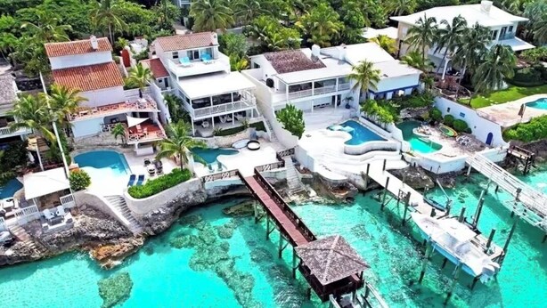 An aerial view of a resort with multiple buildings, a large swimming pool, and a dock extending into turquoise waters. - Bahamas Beach House (Nassau)