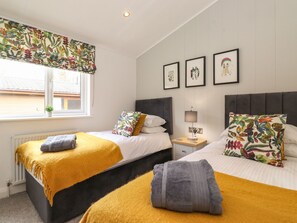 Habitación de hotel con dos camas, una ventana con cortina floral, una mesita de noche con lámpara y cuadros en la pared.