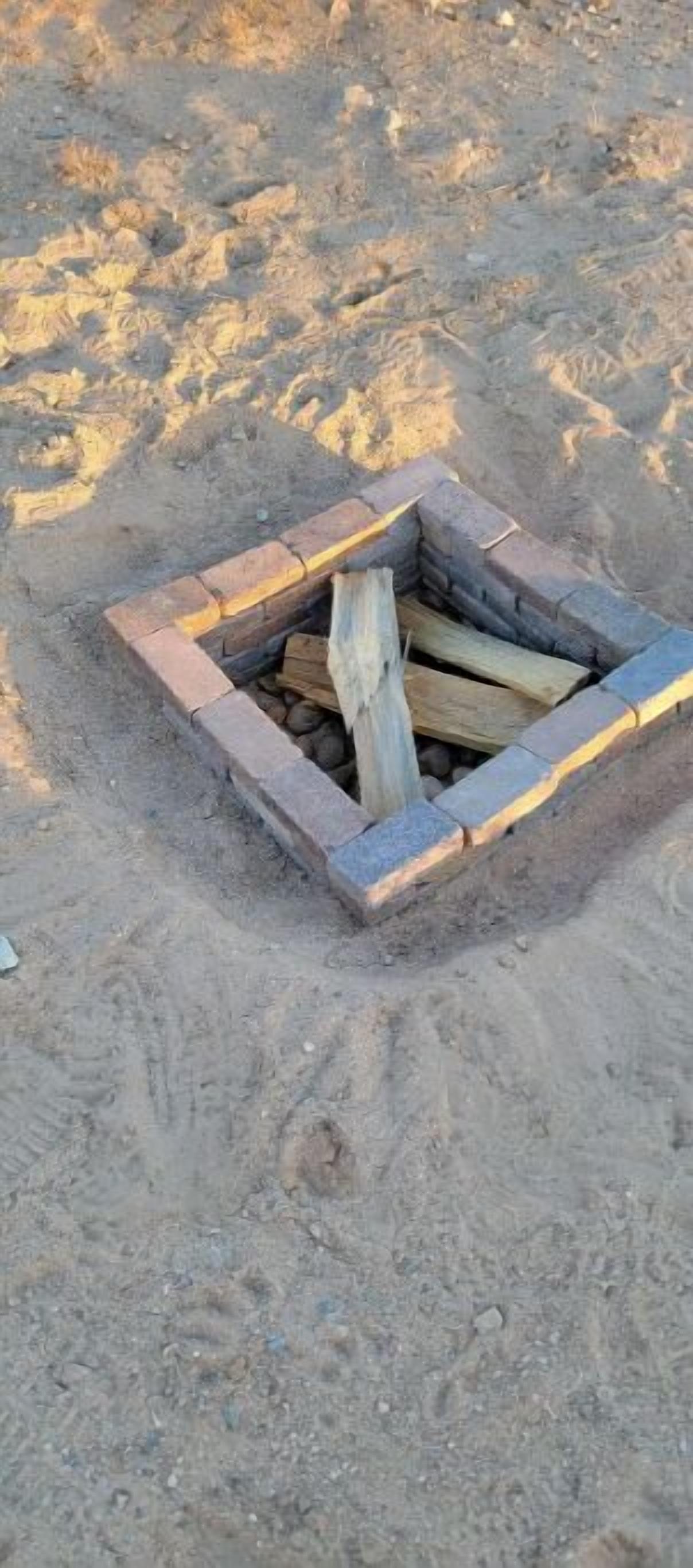 A fire pit with logs and stones, surrounded by a sandy area.