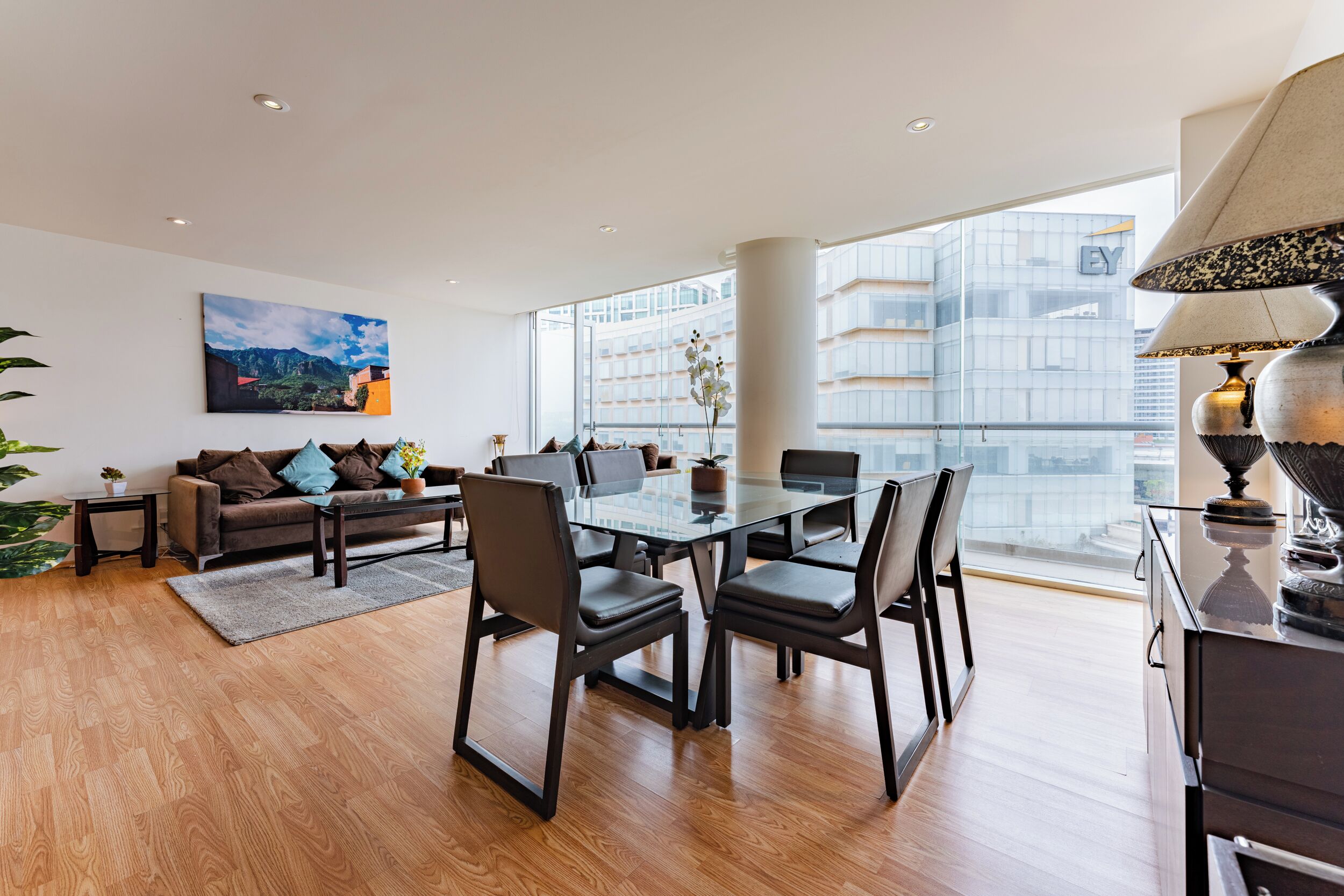 A modern living room with a glass dining table, leather chairs, a sofa, and a large window offering a city view.