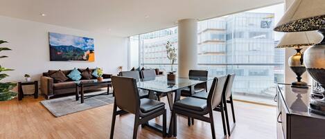 A modern living room with a glass dining table, leather chairs, a sofa, and a large window offering a city view.
