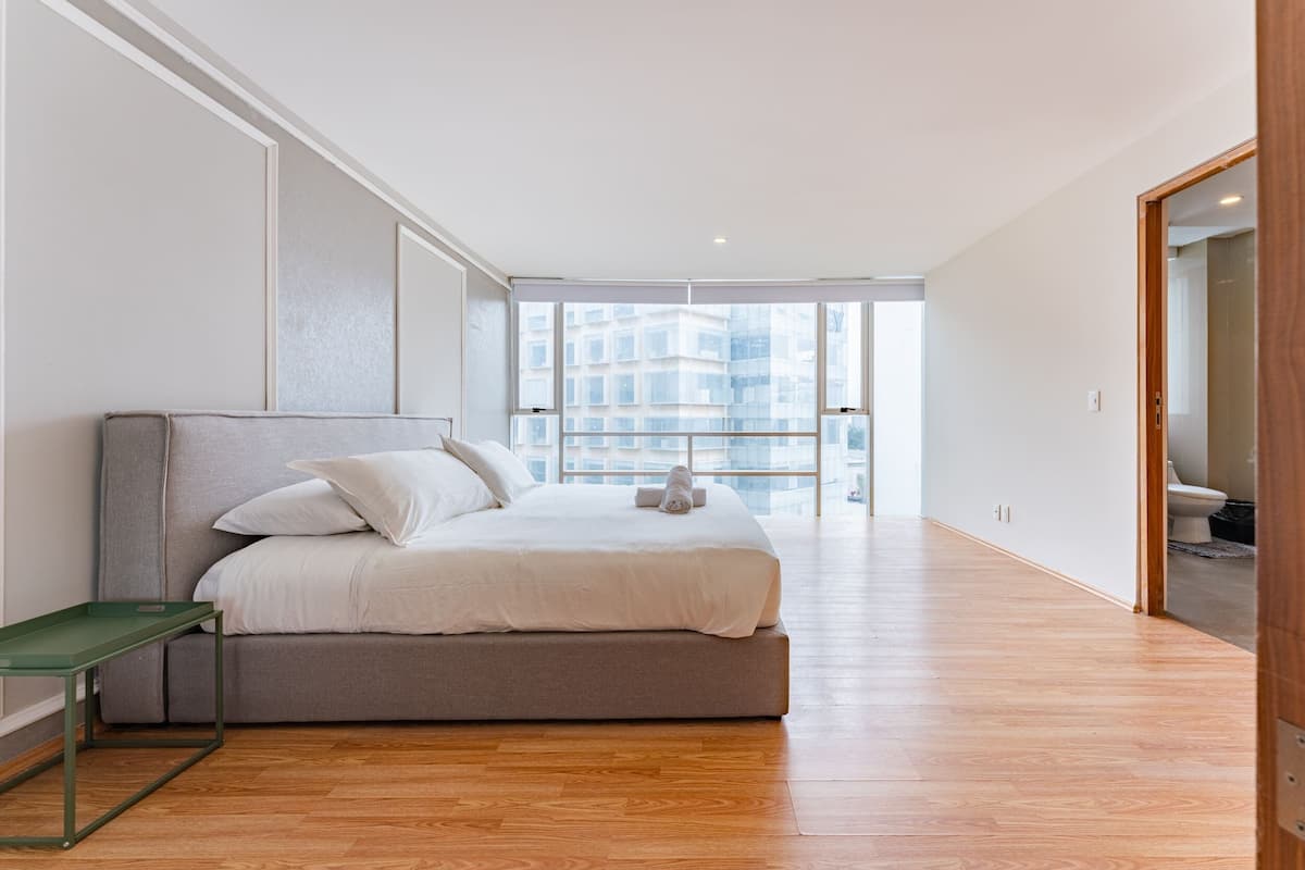 Un dormitorio moderno con una cama, una mesita pequeña y vistas a la ciudad a través de amplios ventanales.