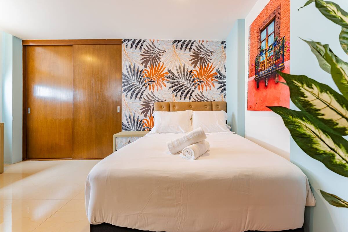 Habitación de hotel con una cama grande, un armario de madera, pared con estampado de hojas tropicales y balcón con pared y ventana roja.