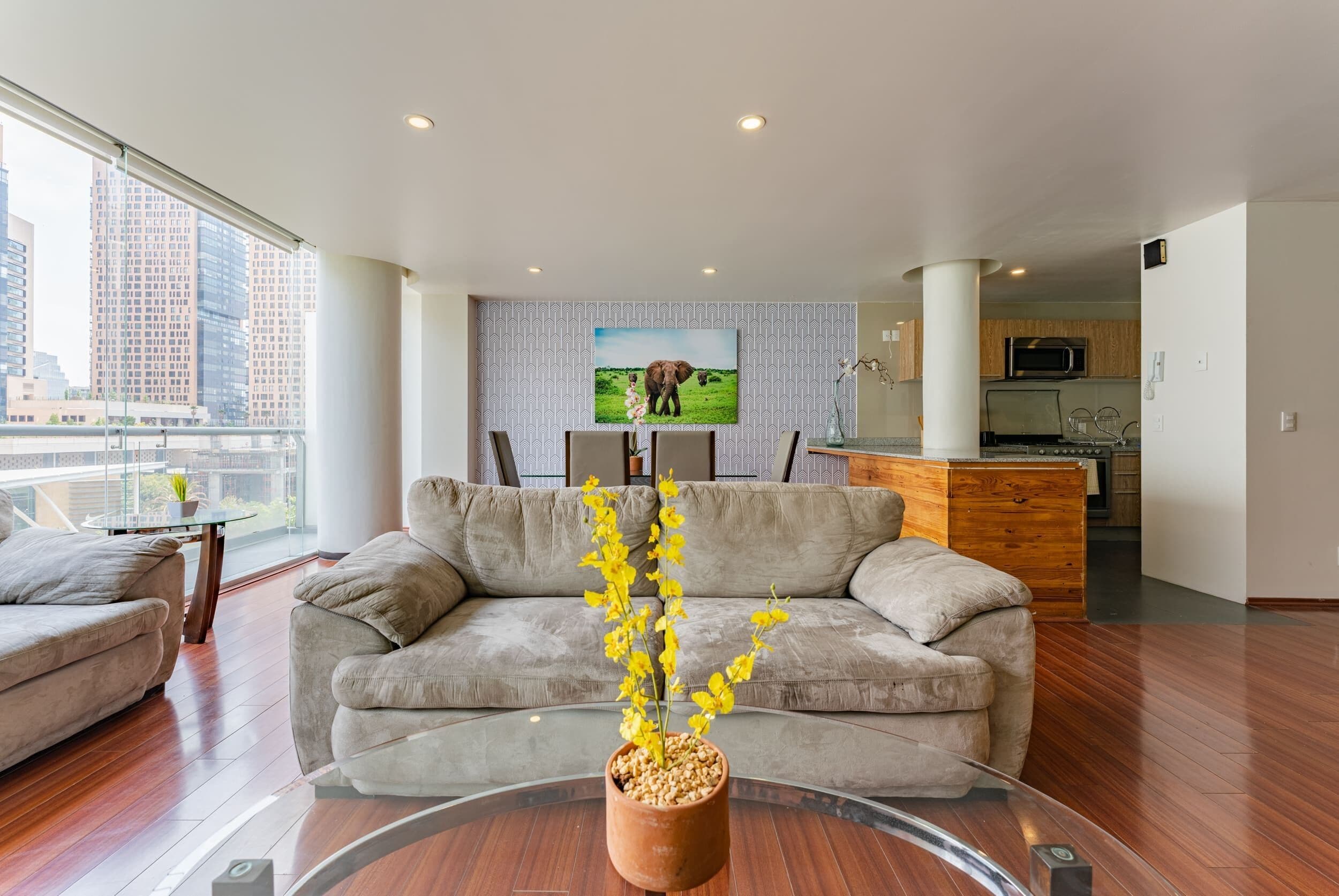 A modern living room with a sofa, a coffee table with a plant, a TV on the wall, and a kitchen area in the background.
