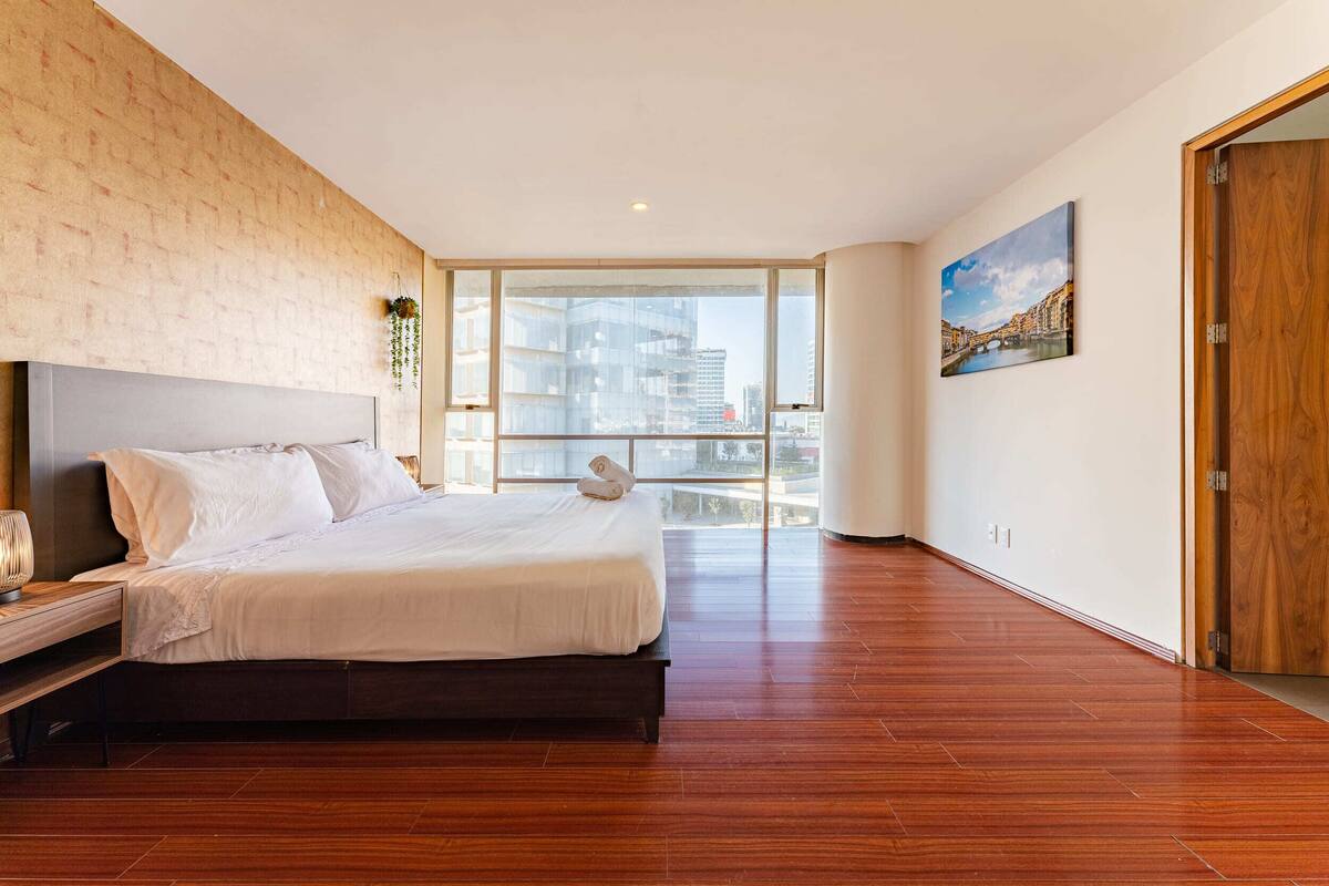 Un dormitorio moderno con una cama grande, un cabecero de madera, una mesita de noche y vistas a un paisaje urbano a través de amplios ventanales.