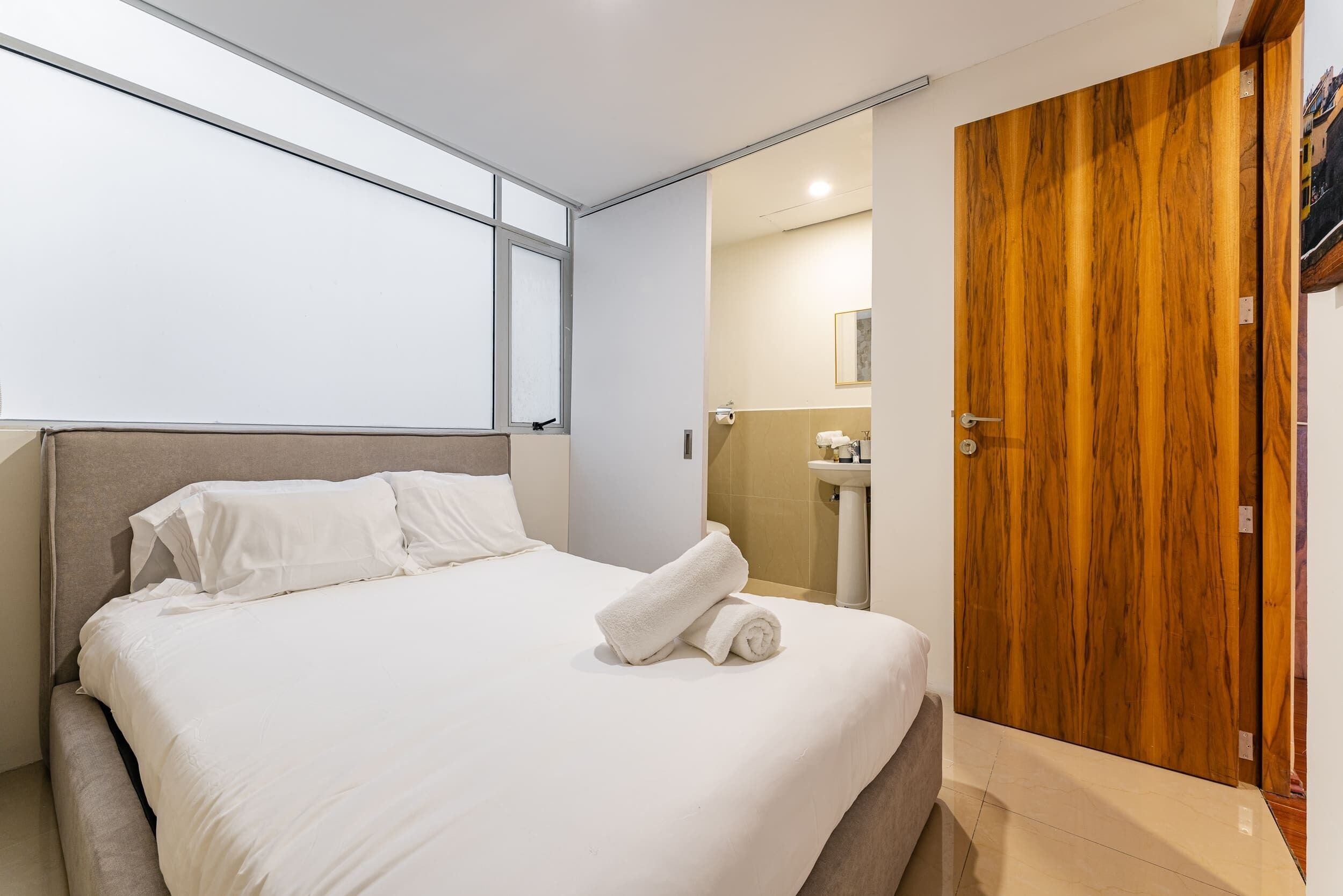 A modern hotel room with a large bed, a wooden wardrobe, a bathroom with a sink, and a window providing natural light.