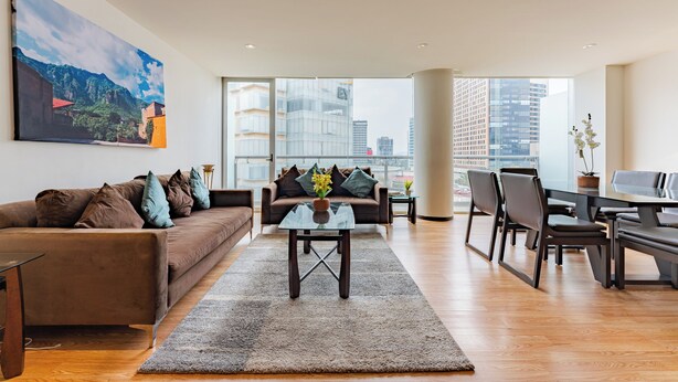A modern living room with a brown sofa, a glass coffee table, and a dining area with a table and chairs. - Antara apartments by VH (Mexico City)