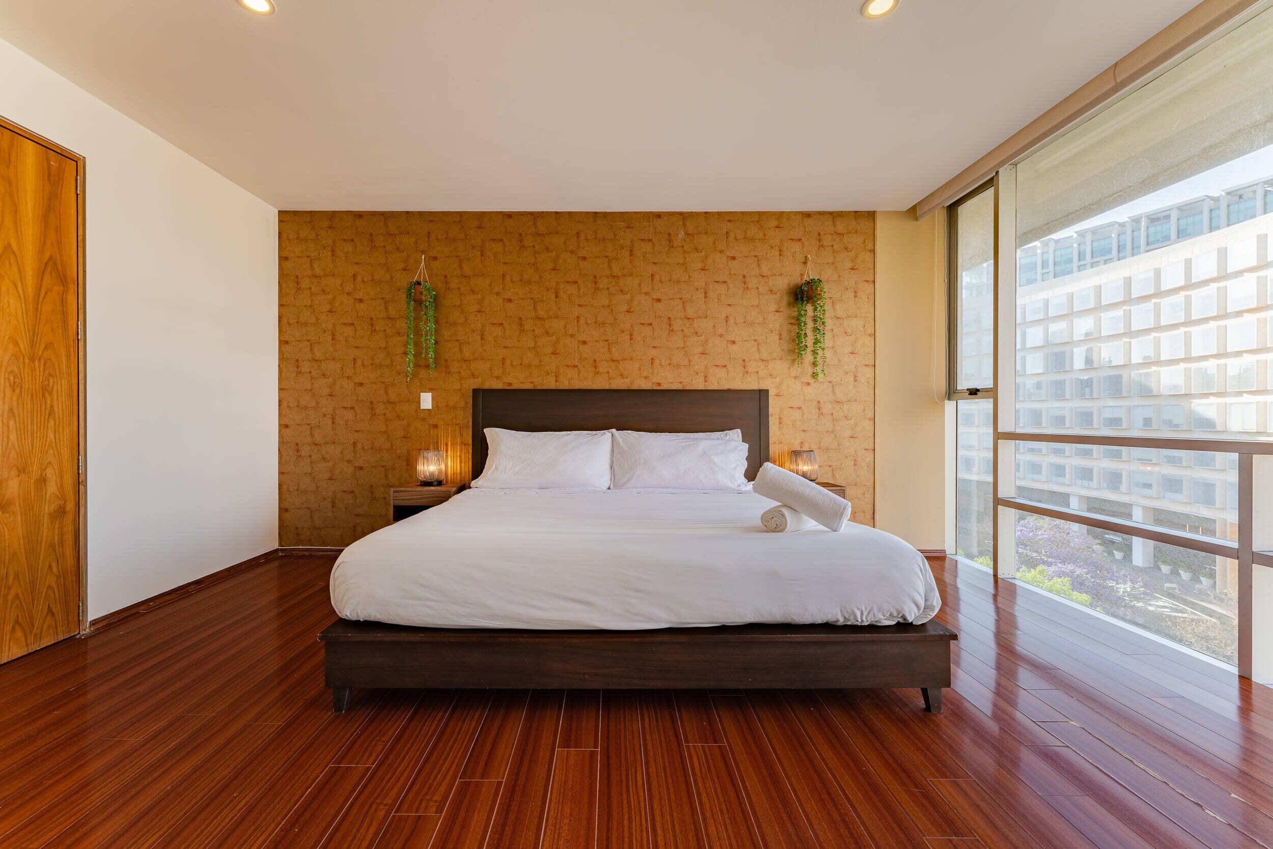 A large bed in a hotel room with a brick wall, wooden floor, and a large window with a view of buildings.