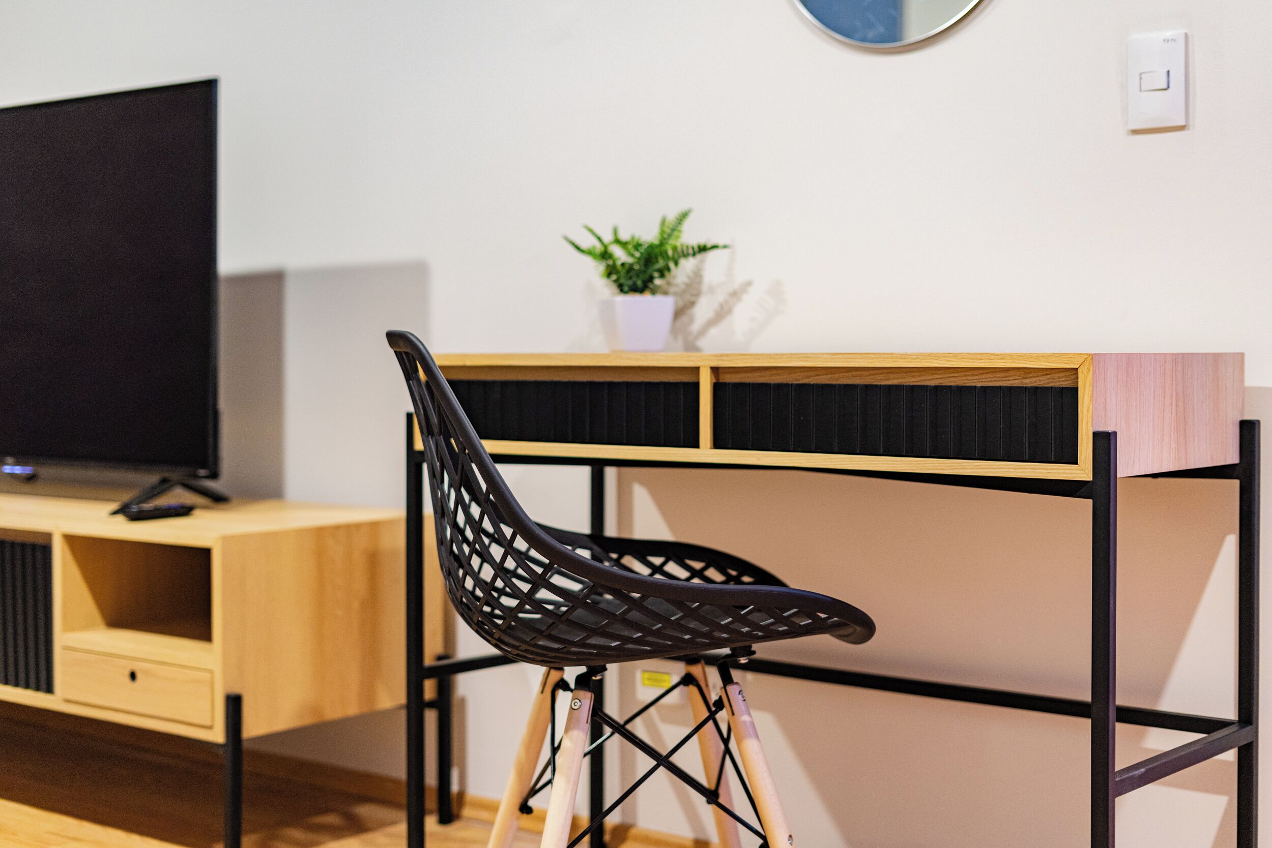 Une chambre moderne avec un bureau en bois, une chaise noire, un téléviseur et une petite plante en pot.