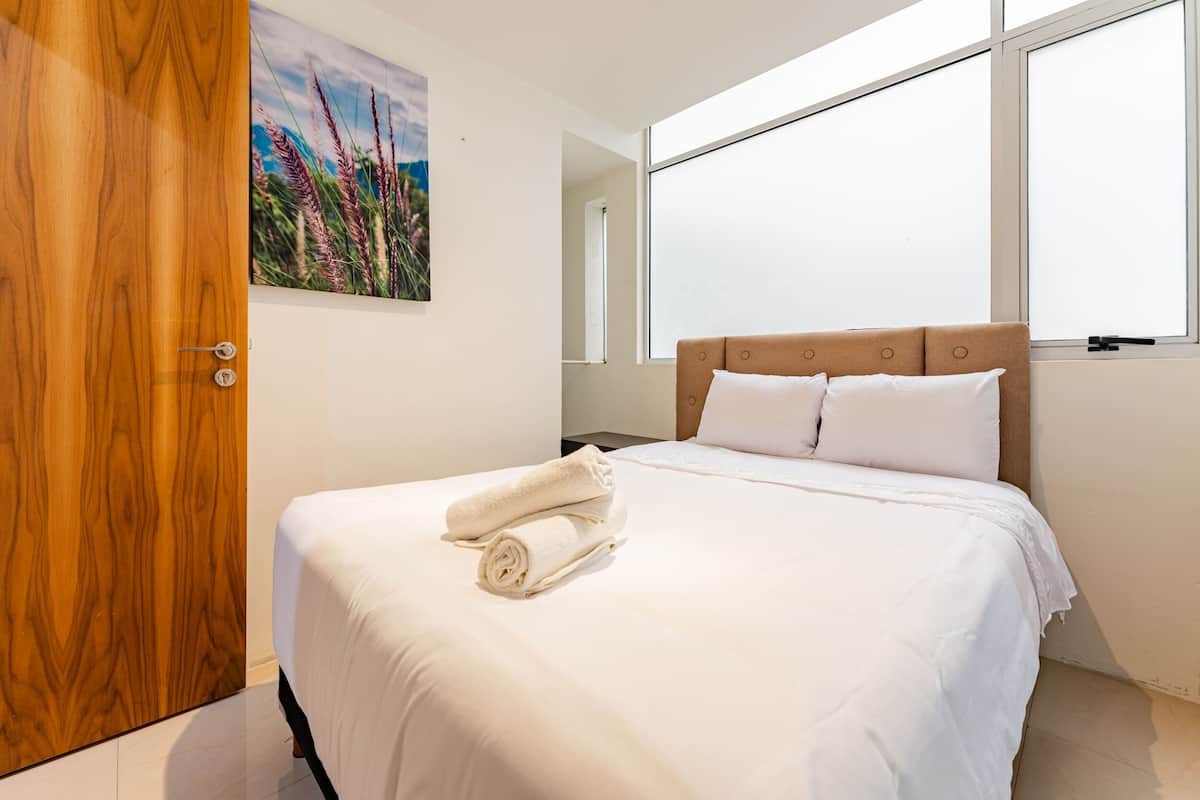 Una cama bien hecha con sábanas blancas y toallas enrolladas, una puerta de madera y un cuadro de hierba alta en la pared.
