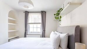 Un dormitorio ordenado con cama, ventana con cortinas, estante con planta y lámpara de noche.