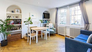 Una sala de estar acogedora con suelo de madera, un sofá azul, una mesa de comedor con sillas, un televisor sobre una estantería blanca y ventanas grandes con cortinas.