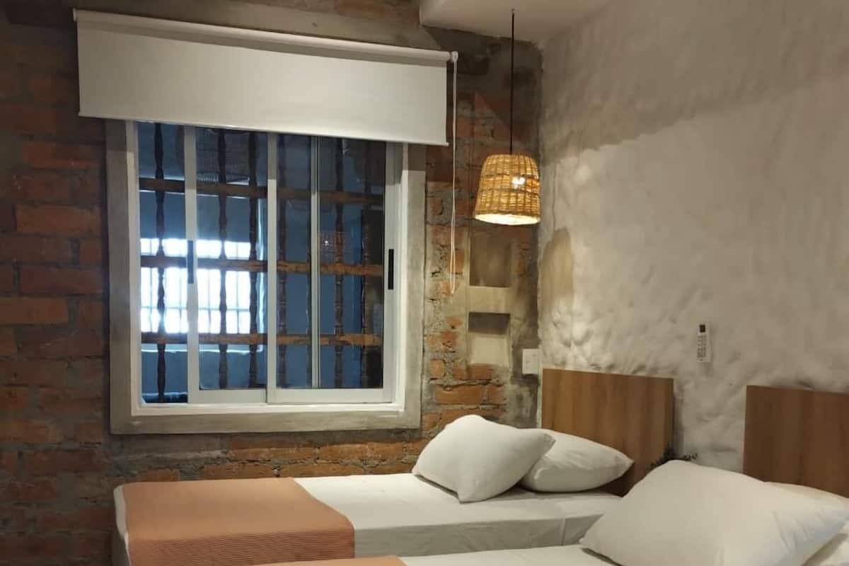 Ein Schlafzimmer mit einem großen Bett, einem Fenster mit Jalousien, einer Pendelleuchte und einer Ziegelwand.