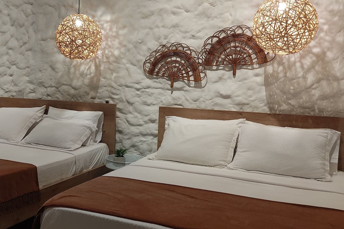 Ein Zimmer mit zwei Betten, einem Holzkopfende, einer weißen, strukturierten Wand und einer Küchenzeile mit Kühlschrank.