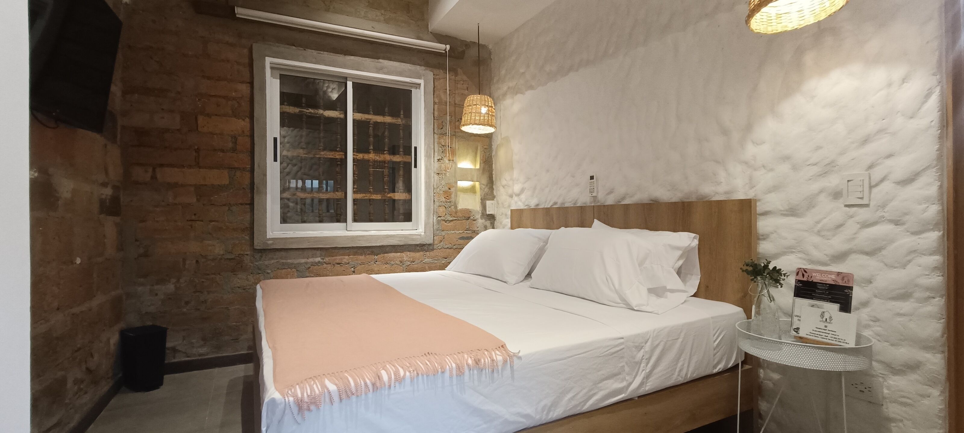 Un dormitorio con pared de ladrillo, un ventanal grande, cabecera de madera, cubrecama blanco y una pequeña mesa con un cartel de bienvenida.