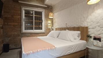Un dormitorio con pared de ladrillo, un ventanal grande, cabecera de madera, cubrecama blanco y una pequeña mesa con un cartel de bienvenida.
