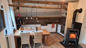 A cozy kitchen with a dining table, wicker chairs, a wooden ceiling, and a wood-burning stove.