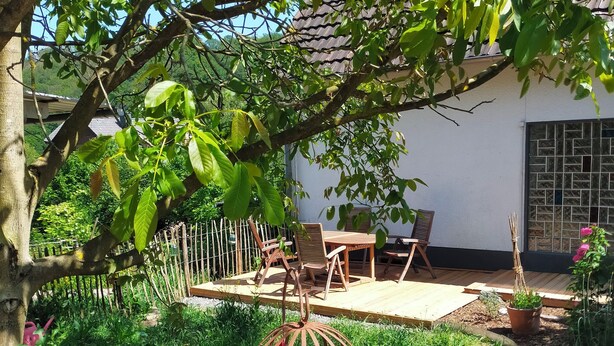 A garden with a wooden table and chairs, a hanging flower basket, and a white building with a tiled window.
