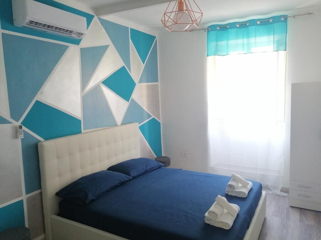 Un dormitorio con un diseño geométrico en la pared, una cama con ropa de cama azul y una ventana con cortinas de tul.