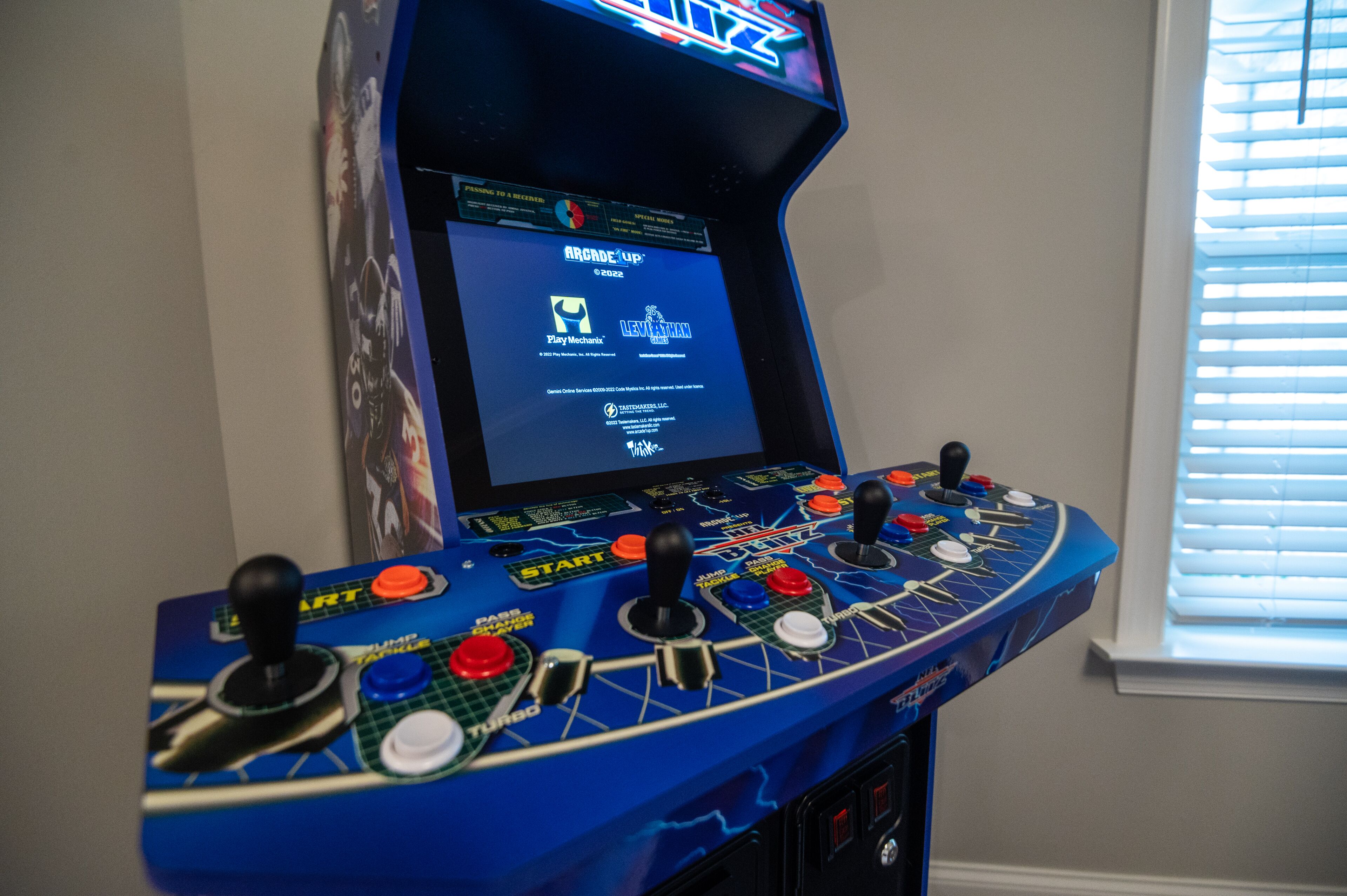 A blue arcade machine with multiple joysticks and buttons, a screen displaying game titles, and a window with blinds in the background.