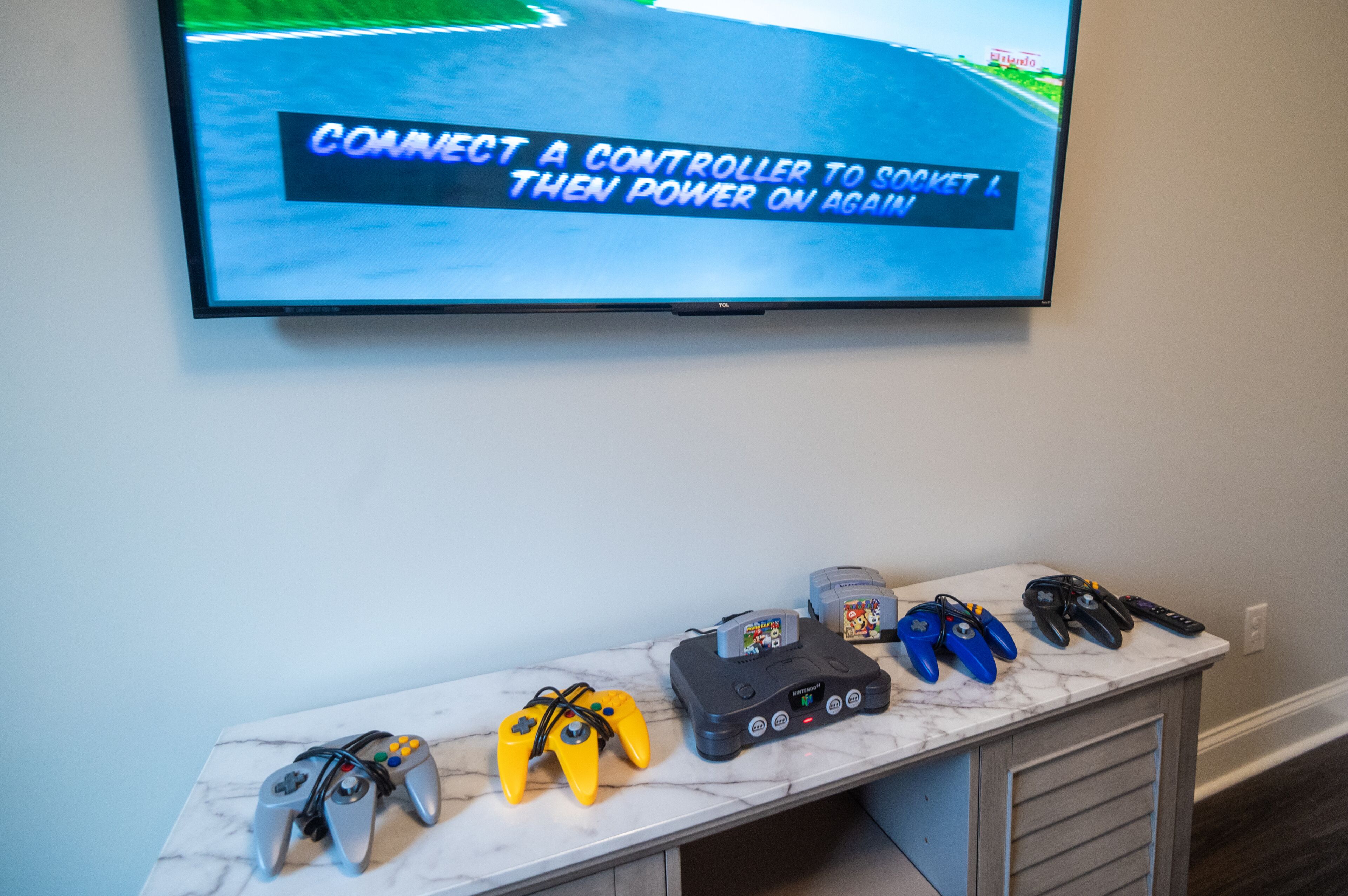 A marble-topped table with various gaming controllers and a television displaying a racing game instruction.