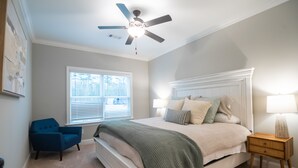 Ein Schlafzimmer mit einem Bett, einem Deckenventilator, einem Fenster, einem Sessel und einem Nachttisch.