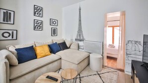 Una sala moderna con un sofá beige, una mesa de centro redonda y un mueble para TV. Hay obras de arte enmarcadas en la pared y una ilustración grande de la Torre Eiffel.