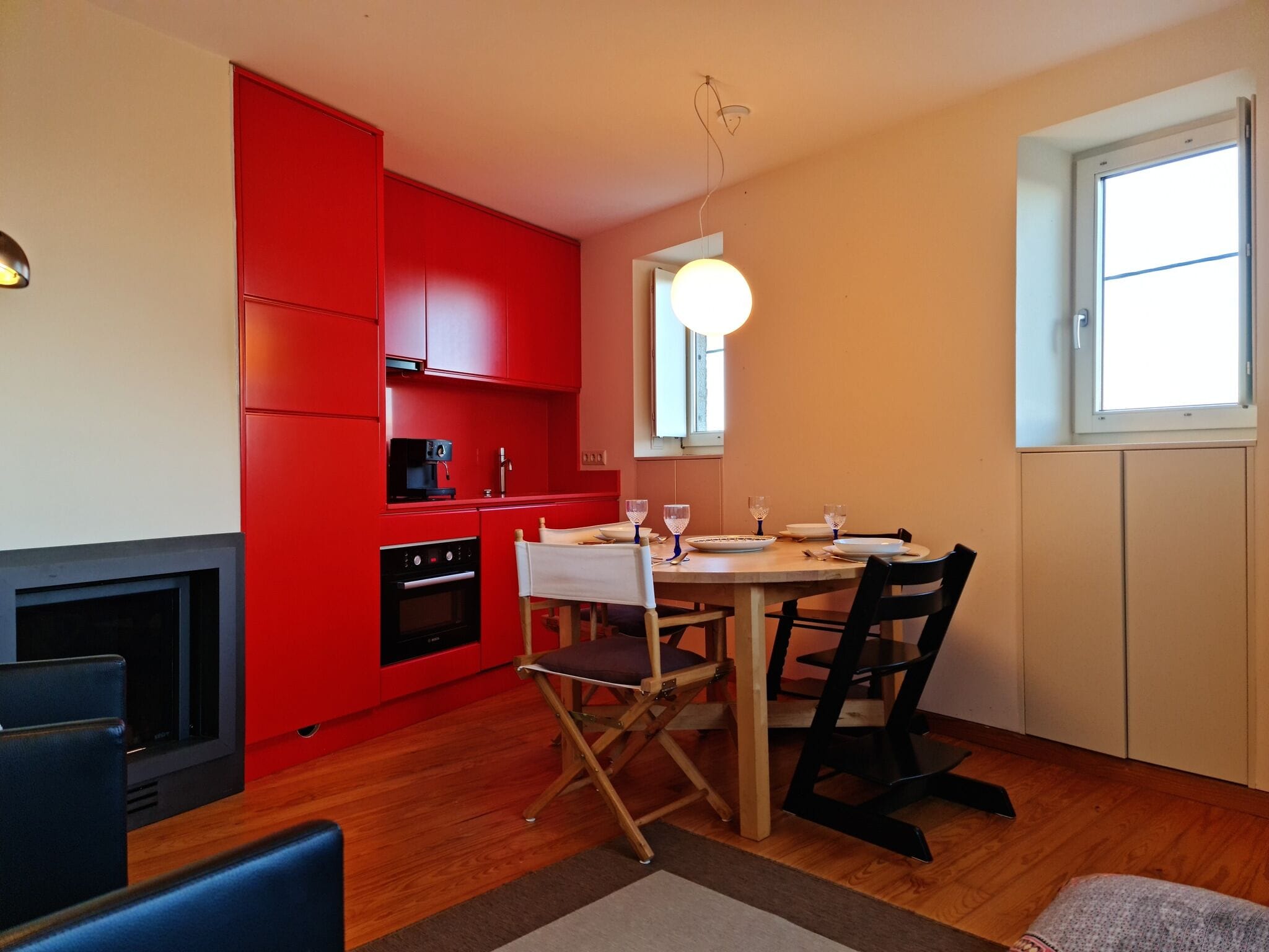Een kleine keuken met rode kastjes, een eettafel met stoelen, een koffiezetapparaat en een groot raam dat natuurlijk licht binnenlaat.