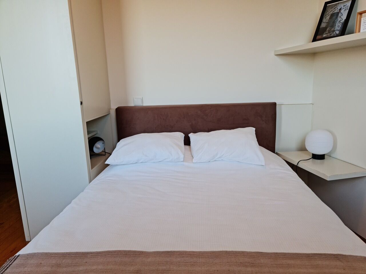 A neatly made bed with white linens and two pillows, a bedside table with a lamp, and a shelf with a framed picture.