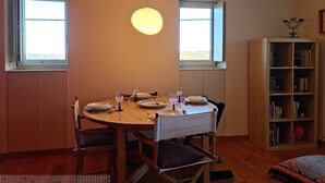 Un coin repas avec une table ronde, des chaises et un luminaire suspendu. Deux fenêtres laissent entrer la lumière naturelle, et une bibliothèque est visible au fond.