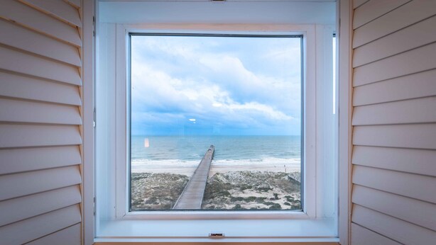 A window with a view of a beach and a wooden pier extending into the ocean. - 779 Amelia by the Sea, Amelia by the Sea 3 Bedroom! Fishing Pier! Oceanfront! (Fernandina Beach)