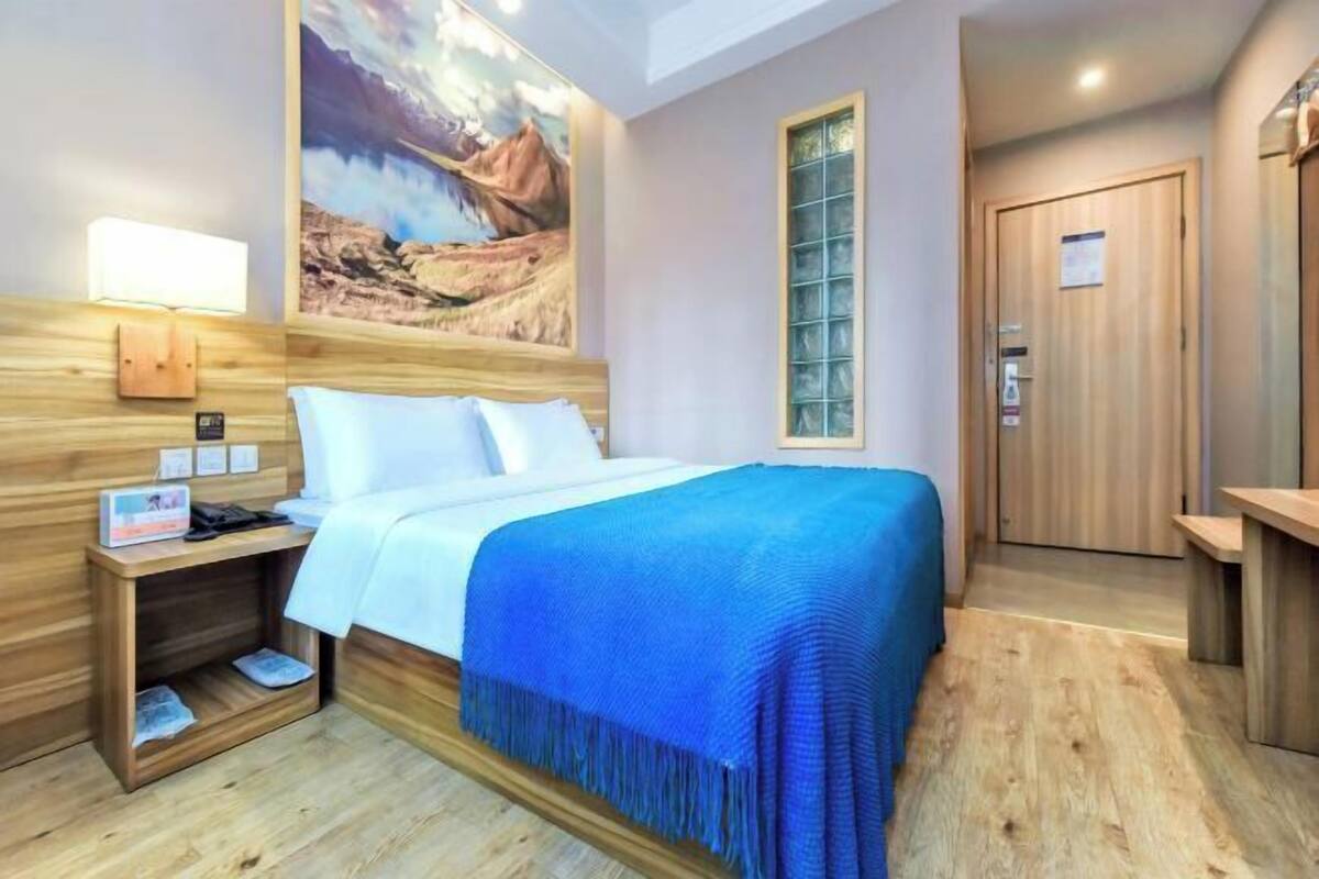 A hotel room with a wooden headboard, a large painting of a landscape, a bedside table with a lamp, a bed with a blue blanket, and a wooden door.