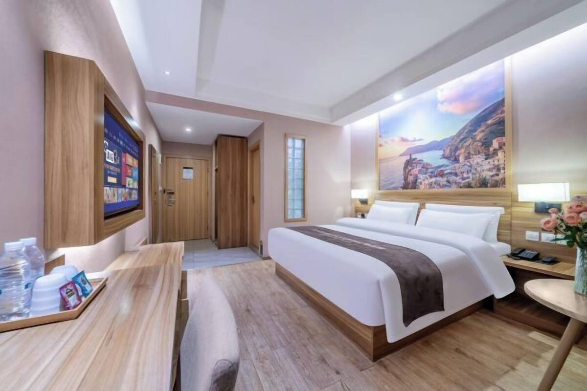A hotel room with a large bed, wooden furniture, a painting of a coastal scene, and a small table with a vase of flowers.