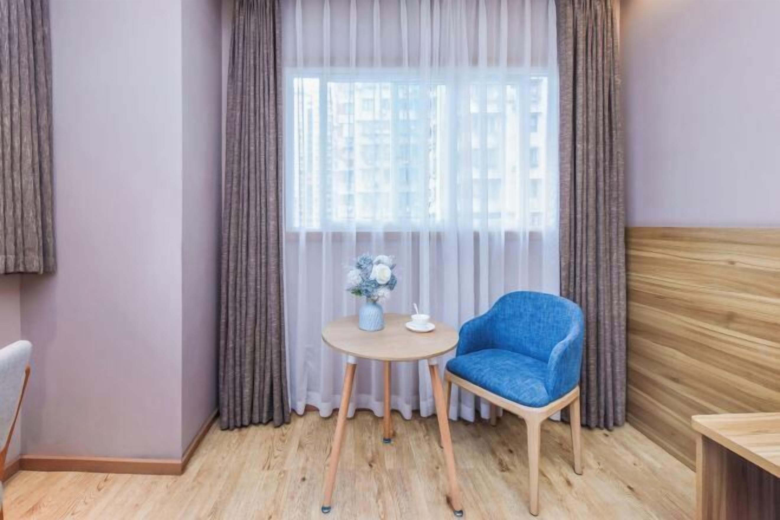 A room with a wooden floor, a blue chair, a small round table with a vase of flowers, and a window with sheer curtains.