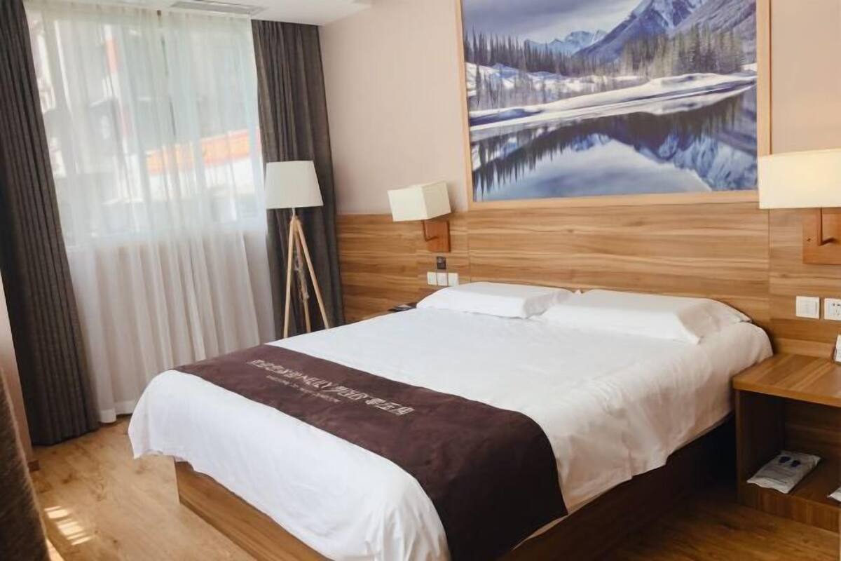 A hotel room with a wooden bed, white bedding, a mountain landscape painting, a lamp, and a bedside table.