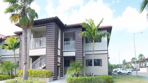 Un edificio residencial de varios pisos con balcones, palmeras y un estacionamiento.