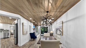 Eine moderne Küche mit weißen Schränken, eine Essecke mit Holztisch und -stühlen sowie eine Decke mit Holzbalken und Einbautenbeleuchtung.