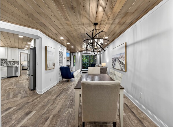 A modern kitchen with white cabinets, a dining area with a wooden table and chairs, and a ceiling with wooden beams and recessed lighting.