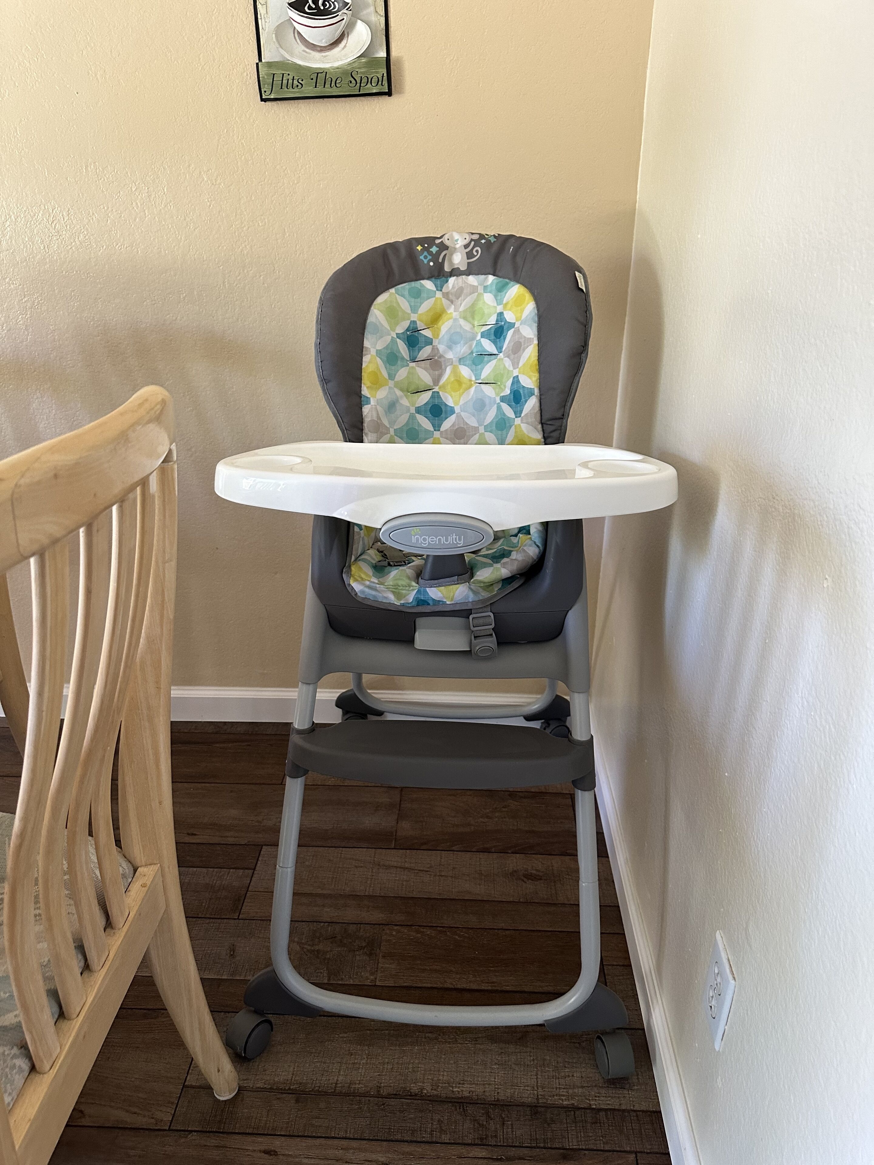 Una silla alta infantil con una mesita, ubicada al lado de una silla de madera en una habitación con suelo de madera y paredes beige.